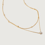 A gold-colored necklace with two layers, featuring tiny spheres on the upper strand and a crystal pendant on the lower chain, displayed against a white background.