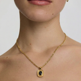 Close-up of a person wearing a gold necklace with a black pendant on a plain background