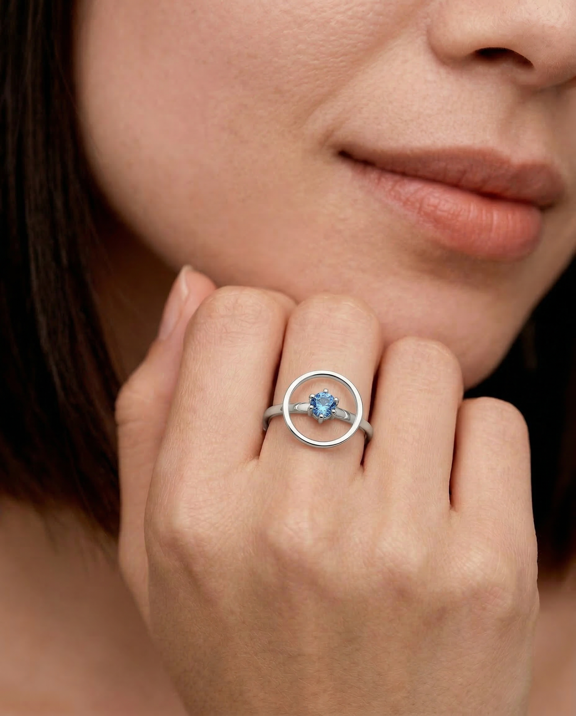 Close-up of a woman's hand wearing a sleek sterling silver ring with a single blue gemstone in the center, set within a circular frame.