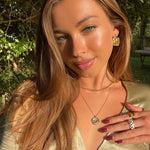 Woman wearing gold jewelry outdoors with greenery in the background