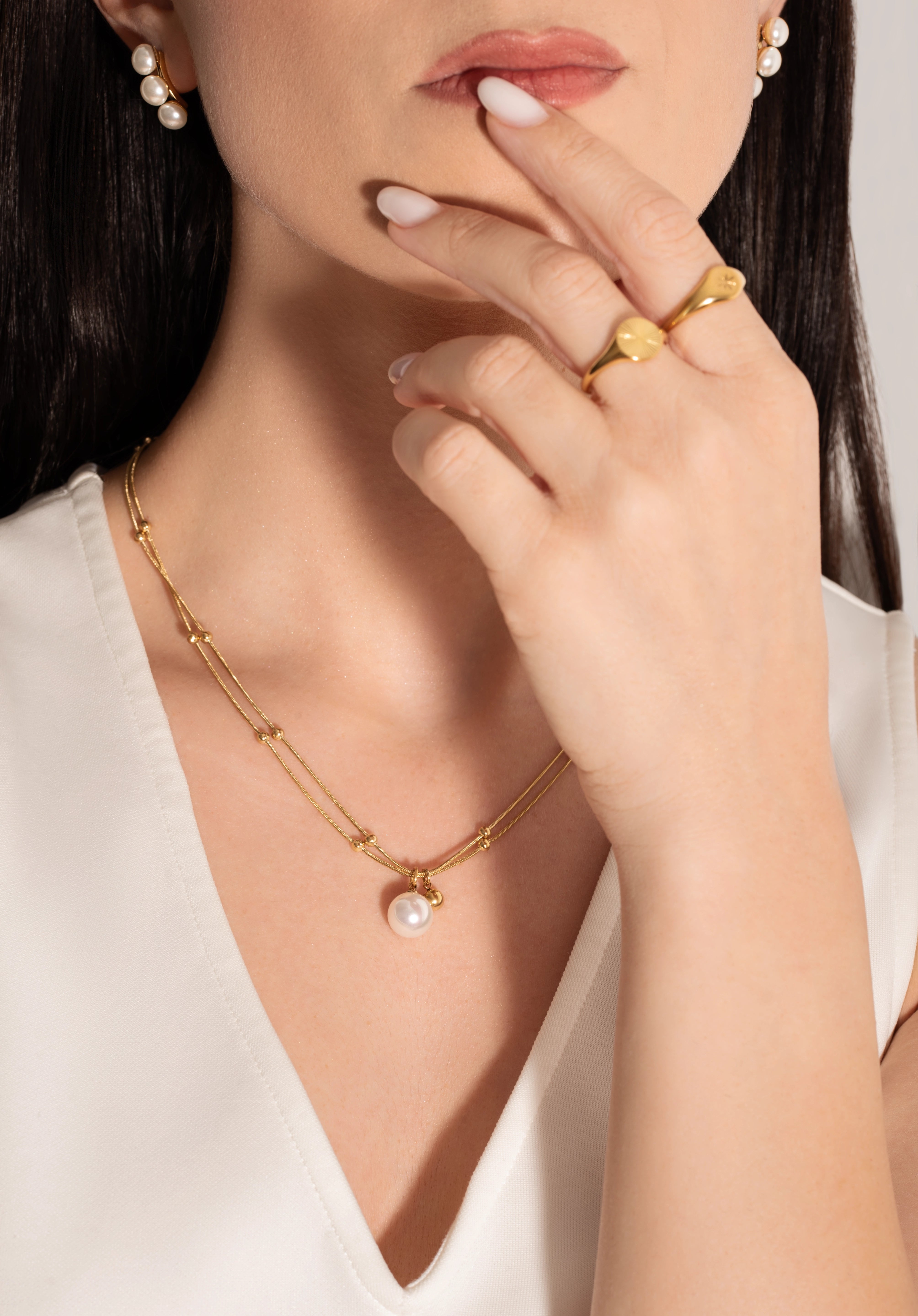 model wearing a gold-plated necklace with two parallel chains, studded with tiny golden spheres and featuring a white pearl pendant.
