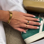 Hand with gold rings on a green sneaker, with a gold handbag in the background.