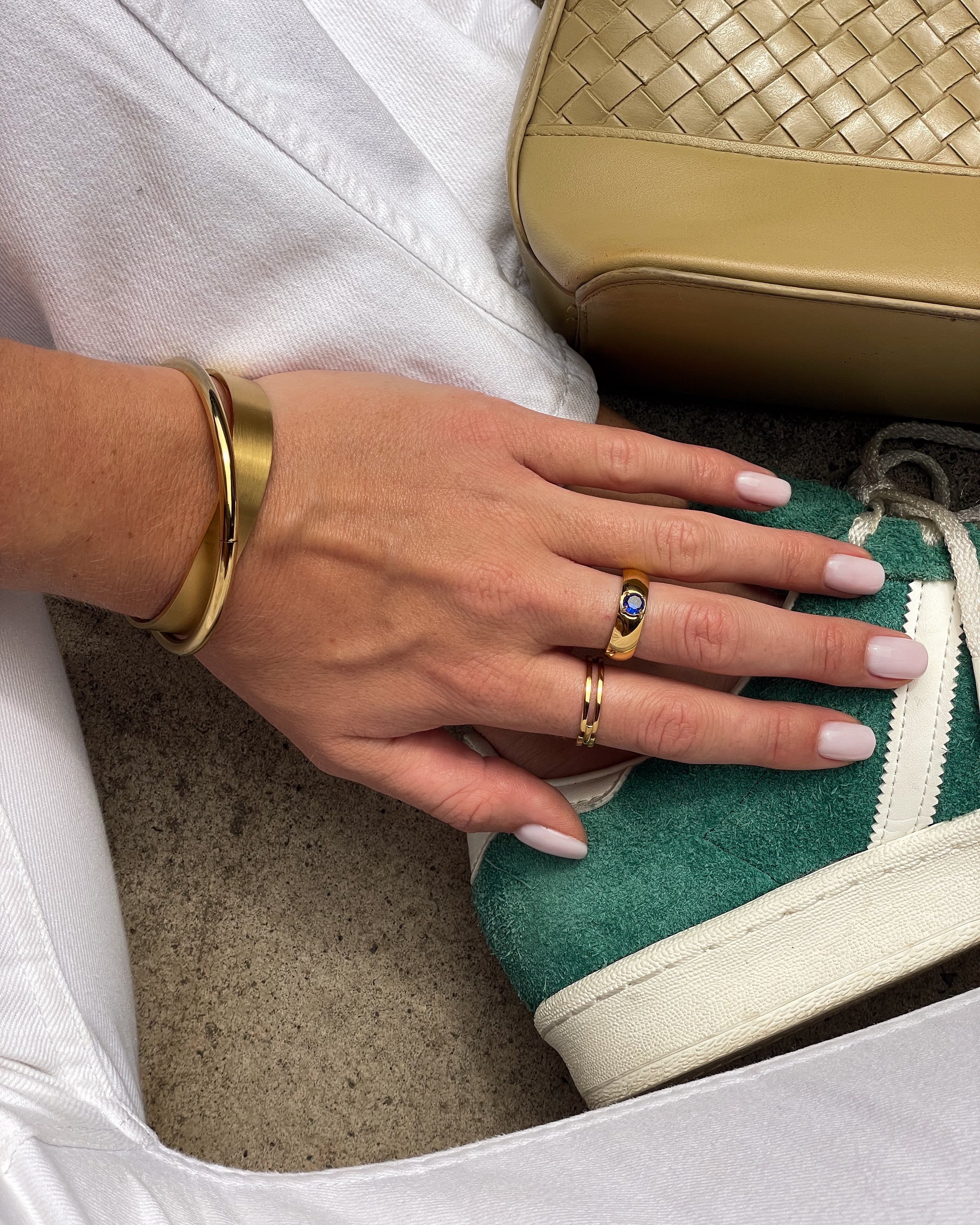 Hand with gold rings on a green sneaker, with a gold handbag in the background.