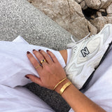 Person wearing a white sneaker with 'N' logo, gold rings, and bracelet on a rocky surface.