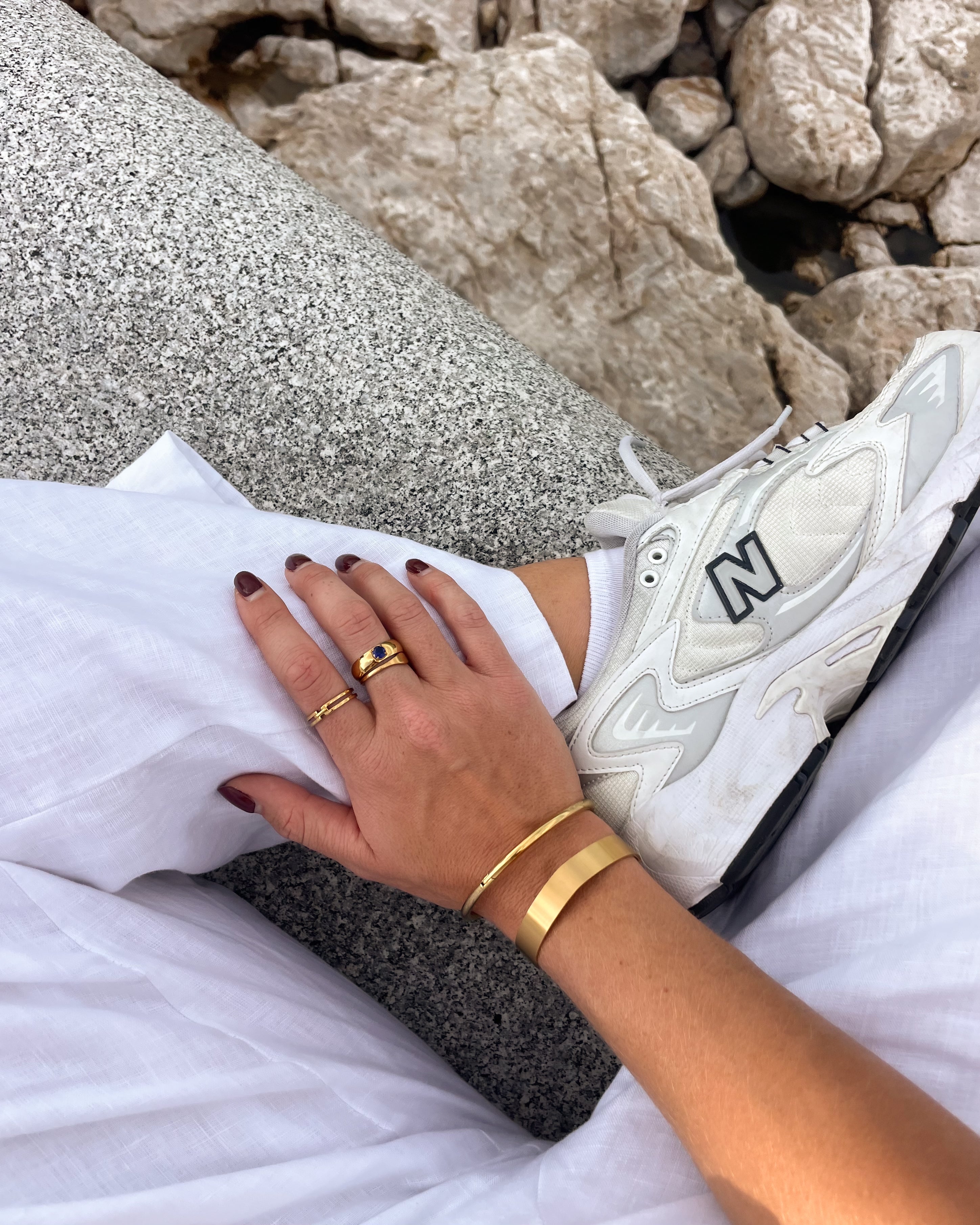 Person wearing a white sneaker with 'N' logo, gold rings, and bracelet on a rocky surface.