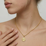 Gold necklace with a circular pendant worn by a person against a neutral background