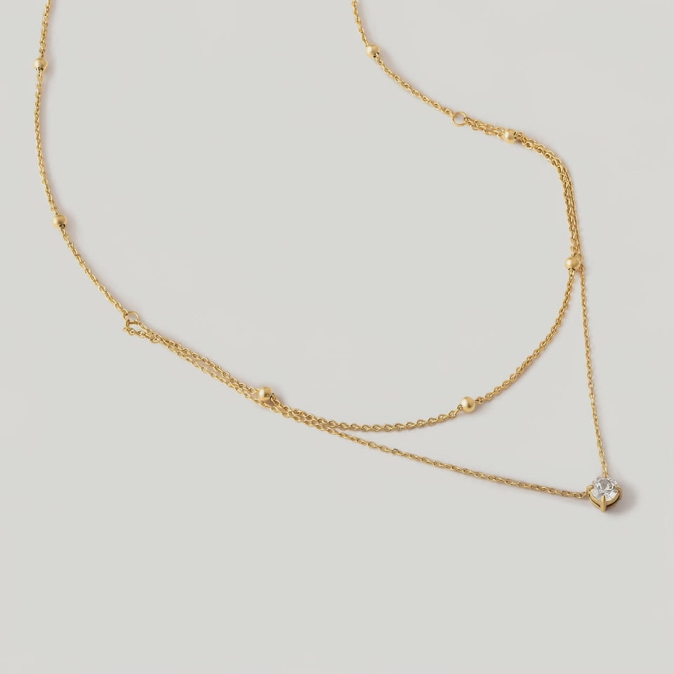 rotating video of a gold-colored necklace with two layers, featuring tiny spheres on the upper strand and a crystal pendant on the lower chain