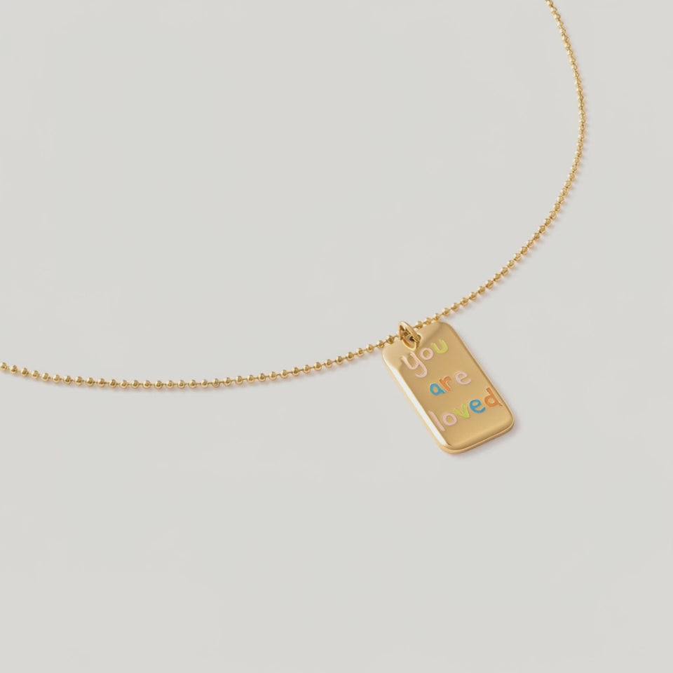video of a a gold plated necklace with a pendant saying "you are loved" in rainbow letters