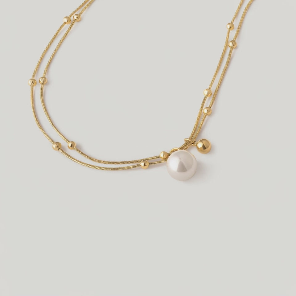 video of a gold-plated necklace with two parallel chains, studded with tiny golden spheres and featuring a white pearl pendant.